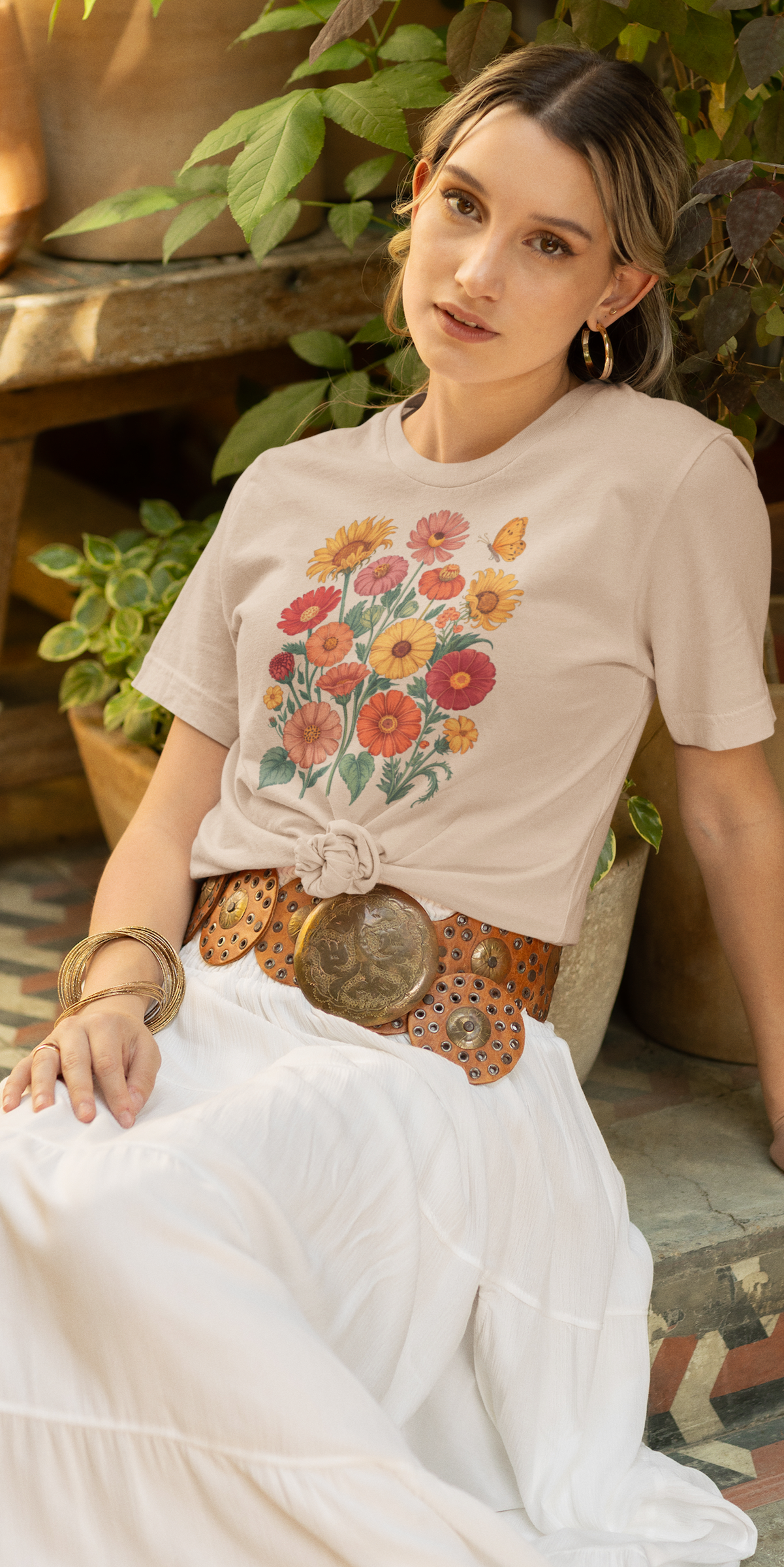 Une femme assise en extérieur entourée de plantes en pot, portant un tee-shirt beige noué sur le côté avec un motif de fleurs colorées et un papillon, associé à une jupe blanche et une ceinture en cuir, pour HappyAndShiny.com.