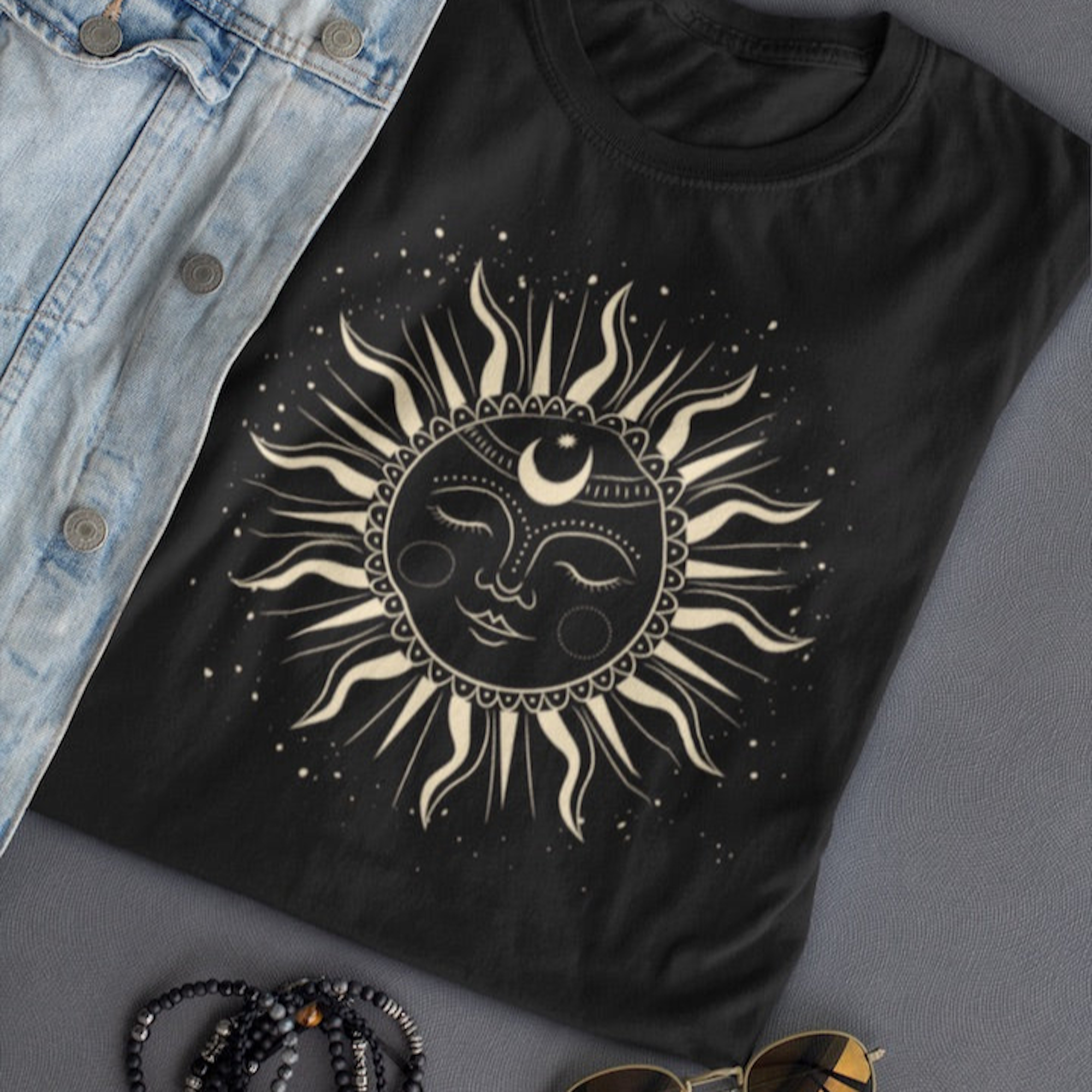 n tee-shirt noir avec un motif de soleil mystique orné de détails géométriques et de rayons, posé à côté d’une veste en denim bleu, de lunettes de soleil et de bracelets, pour HappyAndShiny.com.