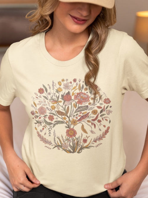 Une femme portant une casquette beige et un tee-shirt blanc orné d’un motif d’arbre floral coloré, assise dans une chambre avec une lampe chaleureuse en arrière-plan, pour HappyAndShiny.com.