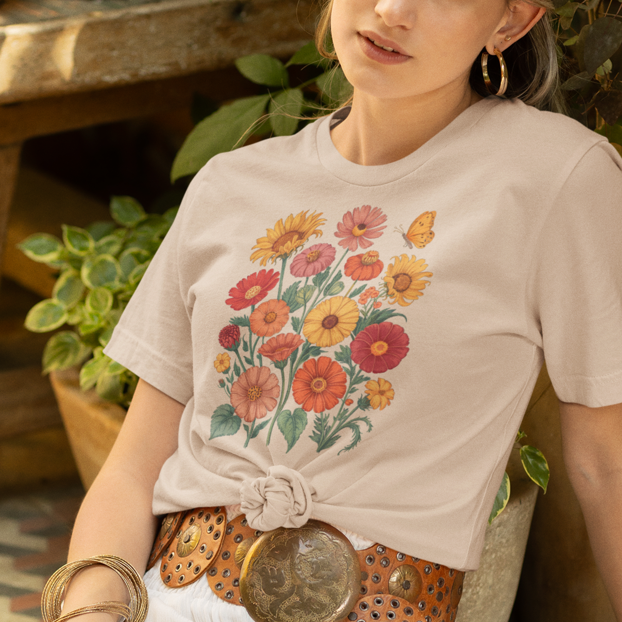 Une femme portant un tee-shirt beige noué sur le côté avec un motif de bouquet floral coloré et un papillon, associé à une jupe blanche, une ceinture en cuir et des bracelets dorés, dans un cadre extérieur verdoyant, pour HappyAndShiny.com.