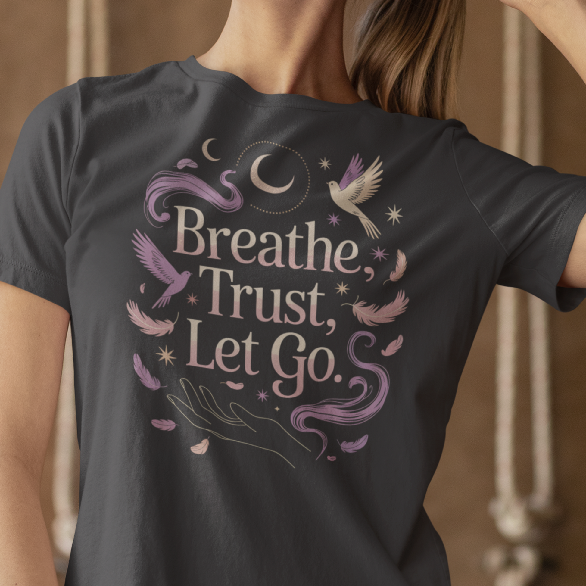 Une femme portant un tee-shirt gris foncé avec l’inscription "Breathe, Trust, Let Go" entourée de plumes, oiseaux et motifs de lune, devant un fond beige, pour HappyAndShiny.com.