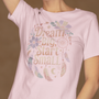 Une femme portant un tee-shirt rose pâle avec l’inscription "Dream Big, Start Small" entourée de plumes, fleurs et motifs de dreamcatcher, devant un fond beige, pour HappyAndShiny.com.