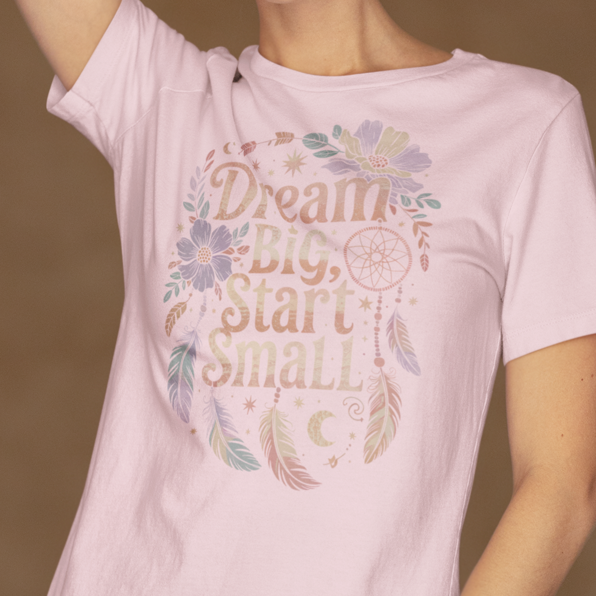 Une femme portant un tee-shirt rose pâle avec l’inscription "Dream Big, Start Small" entourée de plumes, fleurs et motifs de dreamcatcher, devant un fond beige, pour HappyAndShiny.com.
