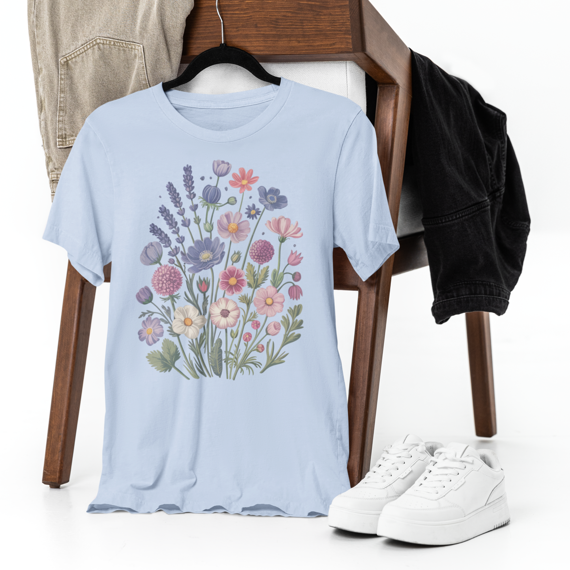 Un tee-shirt bleu clair avec un motif de fleurs sauvages colorées, suspendu sur une chaise en bois avec un jean beige, une veste noire et des sneakers blanches, pour HappyAndShiny.com.