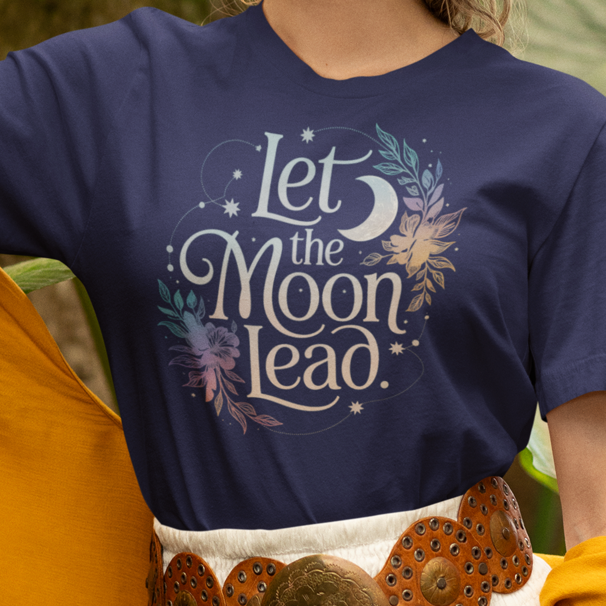 Une femme portant un tee-shirt bleu marine avec l’inscription "Let the Moon Lead" entourée de motifs floraux et de lunes, associé à une jupe blanche et une ceinture en cuir, pour HappyAndShiny.com.