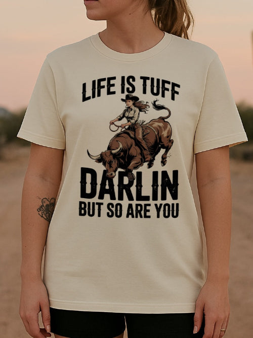 Une femme portant un tee-shirt beige avec un motif de cowgirl montant un taureau et le texte "Life is Tuff Darlin But So Are You", dans un décor désertique avec un cactus, pour HappyAndShiny.com.
