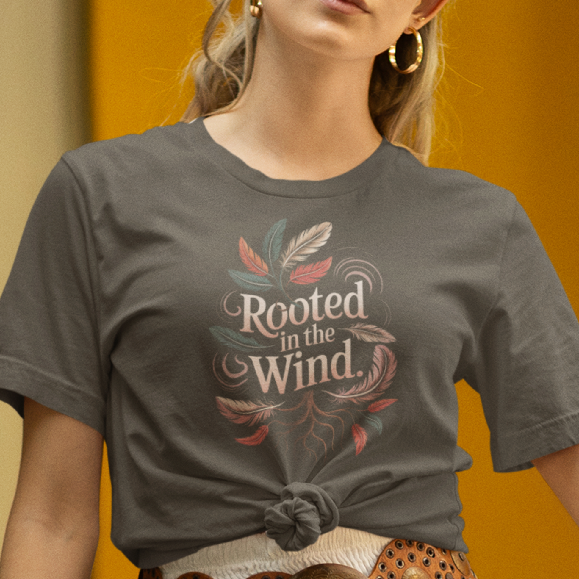 Une femme portant un tee-shirt gris noué sur le côté avec l’inscription "Rooted in the Wind" et des motifs de plumes, associée à une jupe blanche et une ceinture en cuir, devant un fond jaune, pour HappyAndShiny.com.