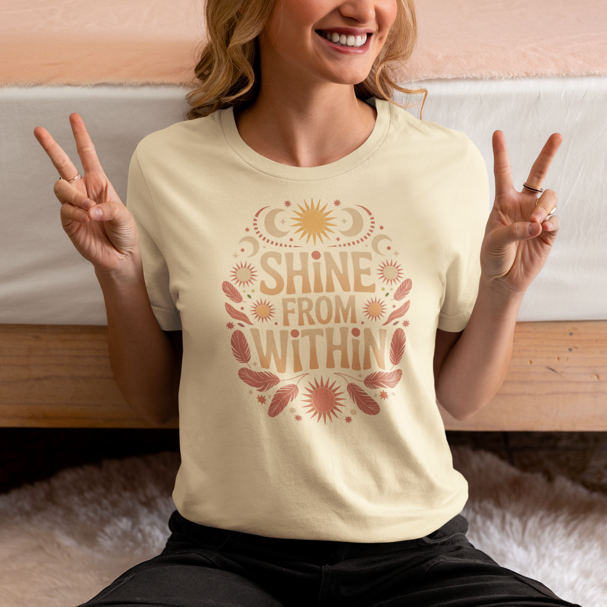 Une femme assise sur un lit, portant un tee-shirt beige avec l’inscription "Shine From Within" entourée de motifs bohèmes comme des plumes, soleils et lunes, faisant un signe de paix, pour HappyAndShiny.com.