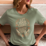 Une femme assise sur un lit, portant un tee-shirt vert avec l’inscription "Let Your Light Guide You" entourée de motifs bohèmes comme des plumes et des astres, pour HappyAndShiny.com.