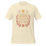 T-shirt unisexe avec un design positif affichant le texte "Shine From Within" (lettres orangées) entouré de motifs célestes (soleils, lunes, étoiles) et plumes beige et rose.