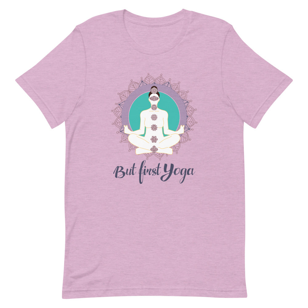 T-shirt rose poudré unisexe avec un design aligné affichant une silhouette méditative blanche en position de lotus, ornée de chakras colorés, entourée d’un mandala violet et turquoise, avec le texte "But First Yoga" en bleu foncé.